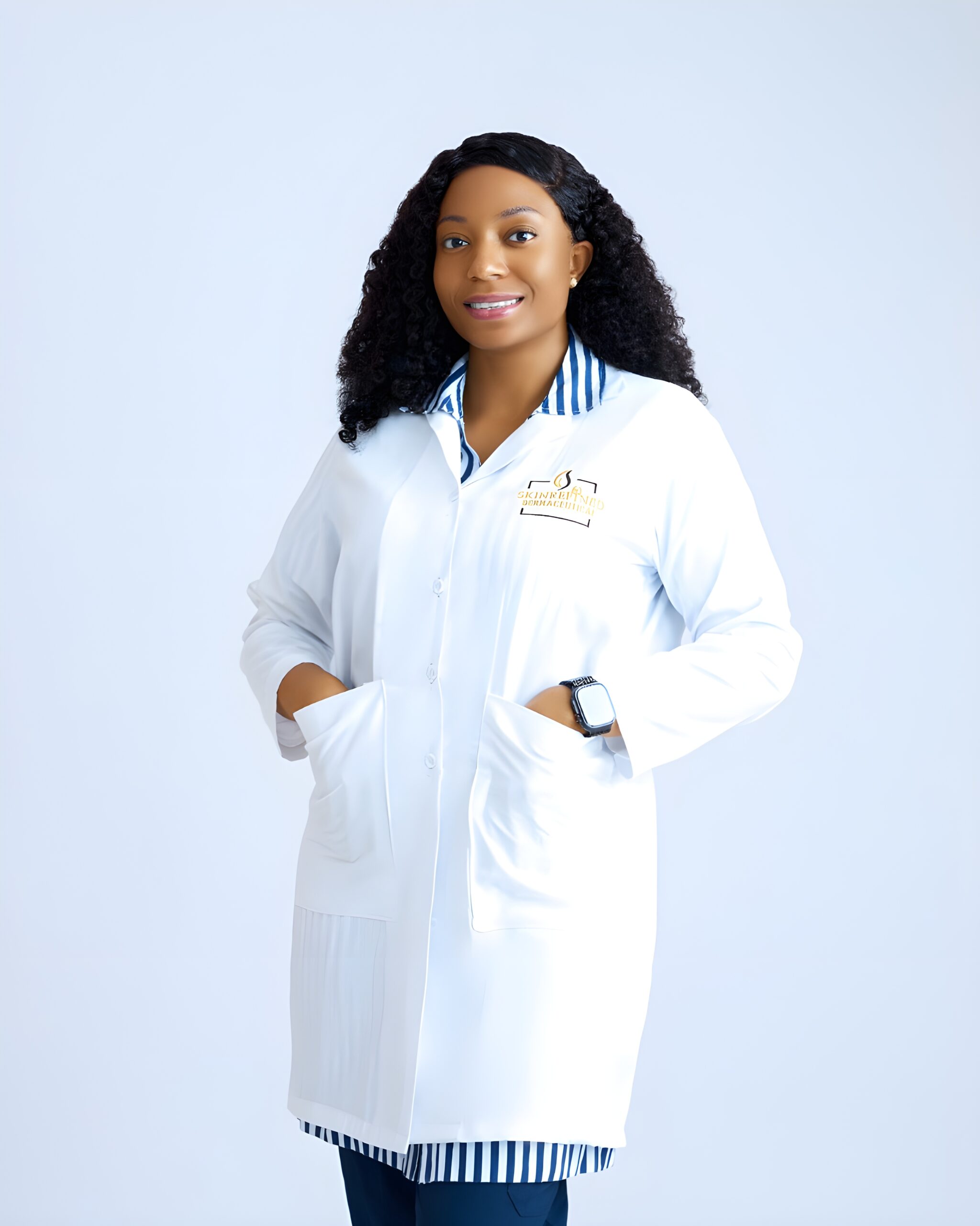 Professional woman wearing a white lab coat over a blue-striped shirt, standing with arms crossed against a pale background.