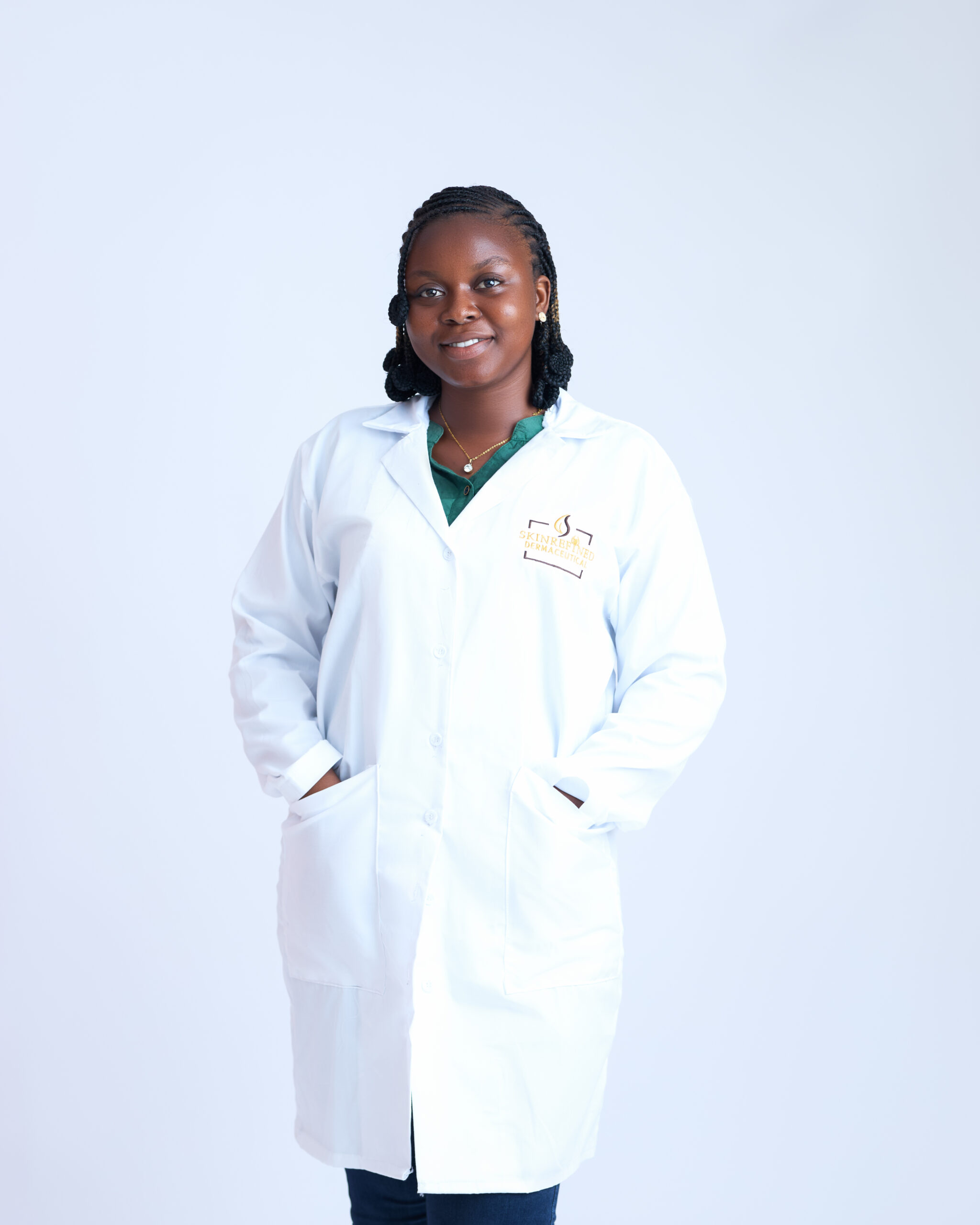 Portrait of a Black woman in a white lab coat over a green top, smiling with hands in pockets in front of a pale blue background.